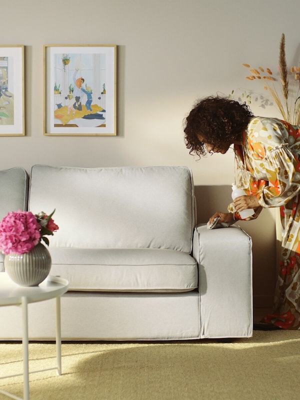 A woman cleaning a light-gray sofa arm with pictures mounted on the wall and a flower pot on a table.
