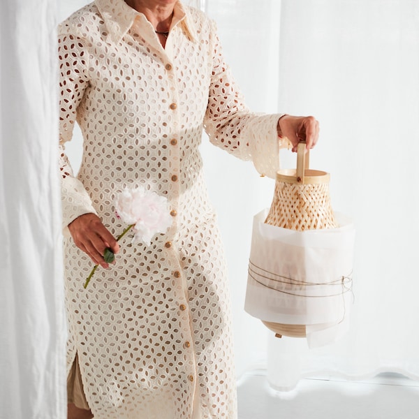 A woman dressed in a beige dress carrying a MISTERHULT table lamp in woven bamboo that is partially wrapped in paper.