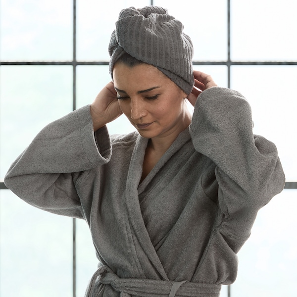 A woman standing in front of a large window wearing a grey ROCKÅN bath robe and a towel wrapped around the top of her head.