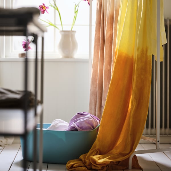 A wooden-floored room with dip-dyed curtains draped over a white MULIG clothes rack and a TORKIS laundry basket next to it.