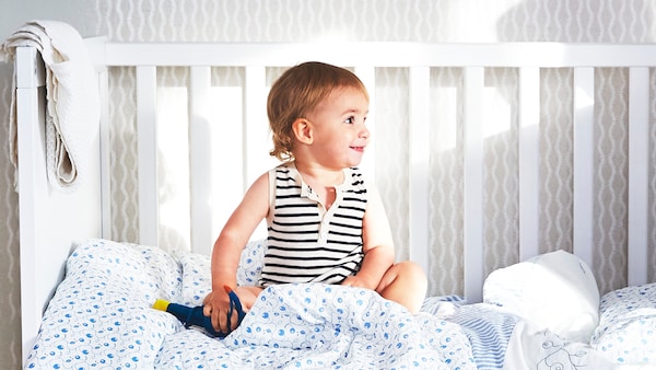 A young child sits in a white SUNDVIK crib with one side removed and GULSPARV bed linen with a blueberry pattern.