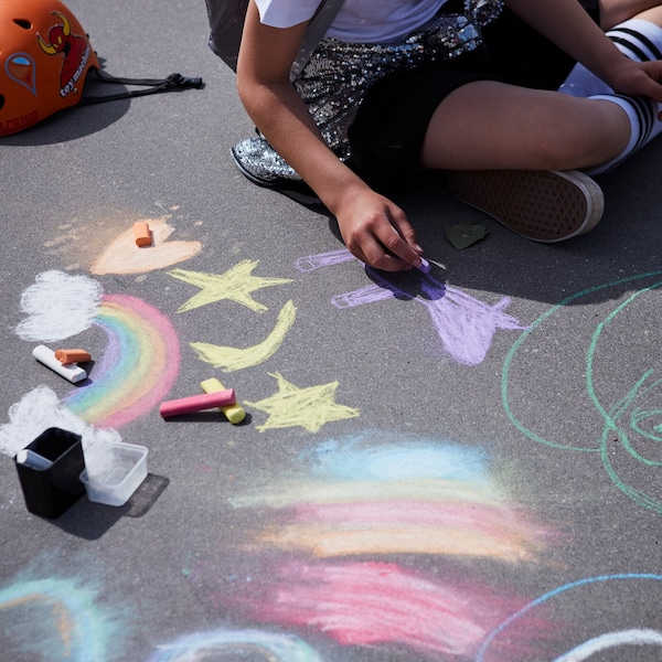 A young girl sits on a concrete surface on a sunny day and draws rainbows and shapes with colored MÅLA chalks.