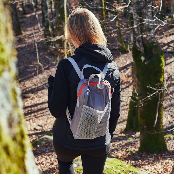 A young person walks beneath a sunlit, sparsely forested hillside, wearing a light-grey PIVRING backpack.