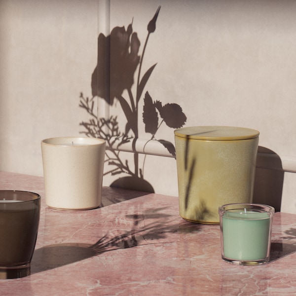ADLAD, HEDERSAM and ENSTAKA scented candles in jars stand on a pink marbled surface with flower-shaped shadows.