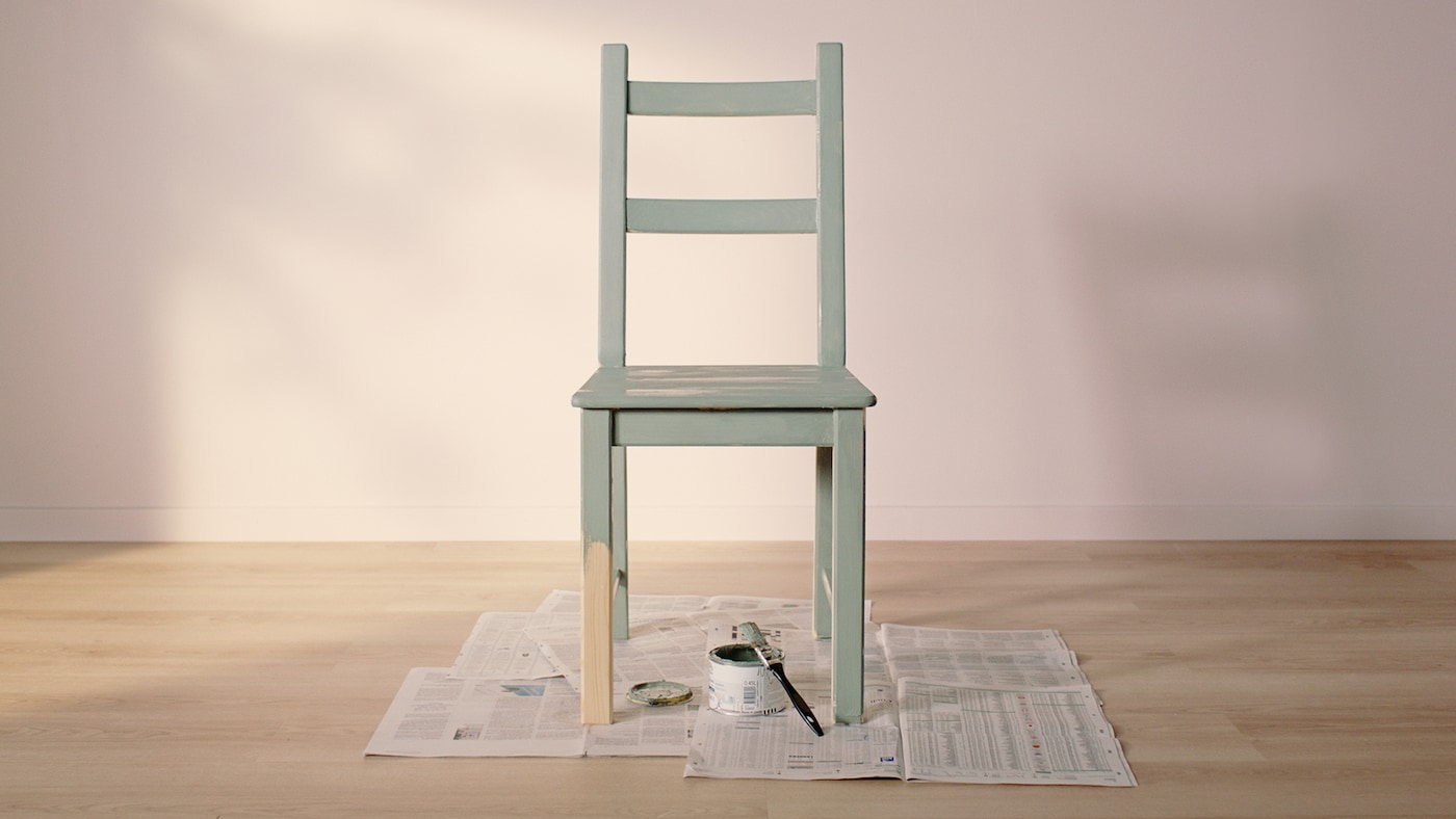 An IVAR pine chair, painted green apart from half of one leg, stands on newspaper with a tin of paint and a paintbrush.