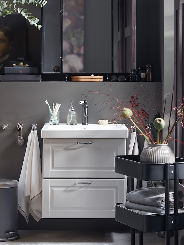 An overall grey bathroom with a sink with bathroom articles on it, a black cart and many dried decorative flowers.