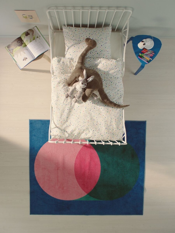 Bird's eye view of a white metal kids bed with a dinosaur and rabbit soft toy, and a blue rug with pink and green circles.