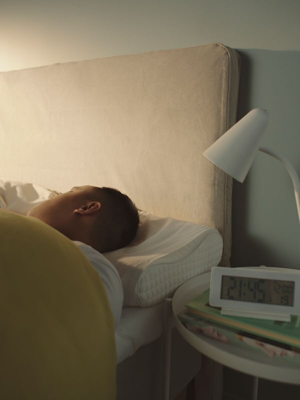 Close-up of a person’s head lying on an ergonomic pillow in bed with a white table lamp and an alarm clock on a side table.