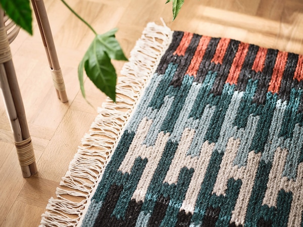 Colorful low pile woven rug on wooden floor next to bamboo plant stand.  