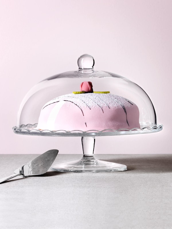 Delicious cake coated with pink icing in a glass pie holder with lid by a cake server utensil. 