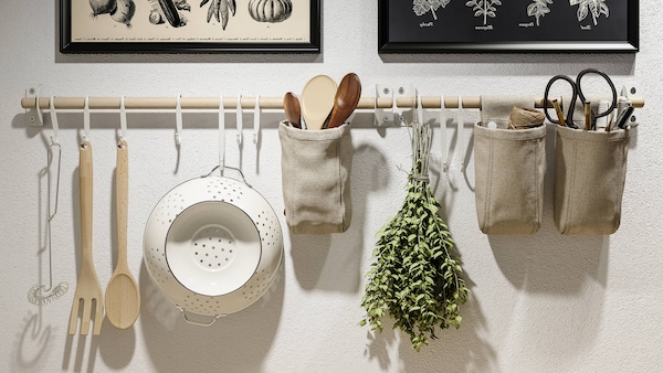 Kitchen utensils and herbs hanging from white metal hooks from a wooden kitchen rack under modern style illustrative prints. 