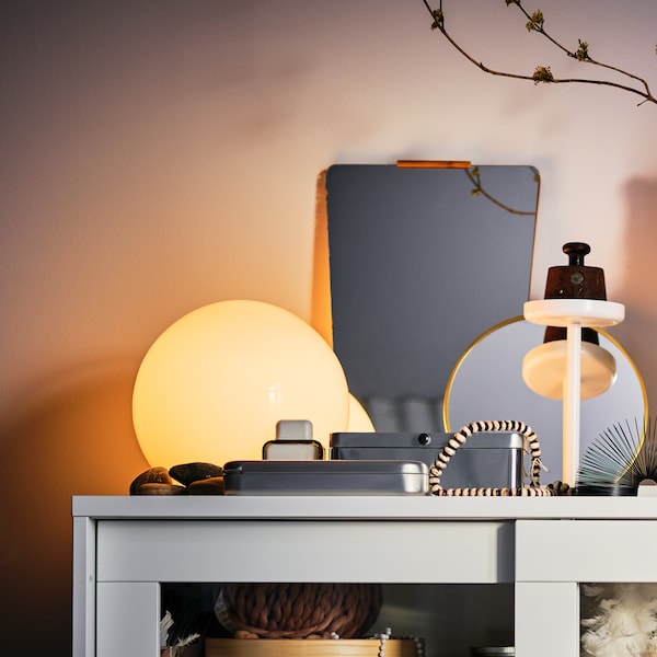 On top of a white SYVDE glass-door cabinet stands a lit, white FADO table lamp amid multiple decorative items.