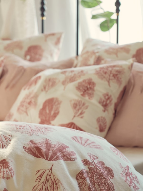 Part of a bed with a duvet and some pillows in VITPYROLA bed linen with a seashell and coral pattern and two pink cushions.