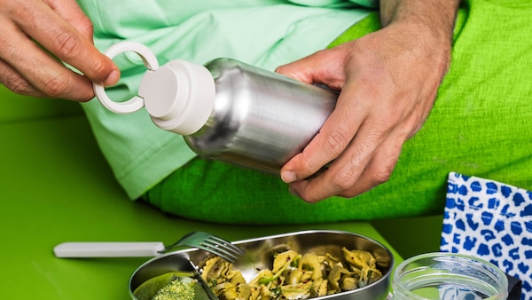 Person in green pants opening a reusable water bottle while eating a meal with reusable containers and utensils. 