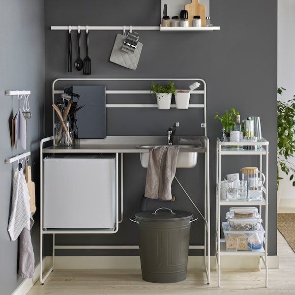 A SUNNERSTA mini-kitchen against a dark grey wall and a kitchen trolley with glasses, plastic containers and jars.