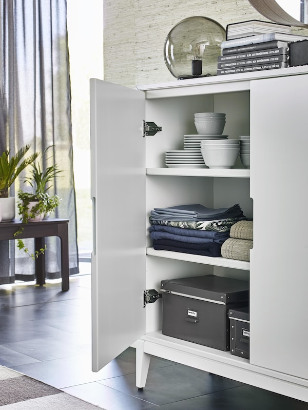 An open REGISSÖR white sideboard cabinet shows stacked plates and bowls, folded tablecloths and linen, and black boxes.