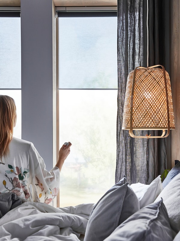 A woman, on a bed near a KNIXHULT pendant lamp, uses a remote control on KADRILJ roller blinds hanging in the windows.