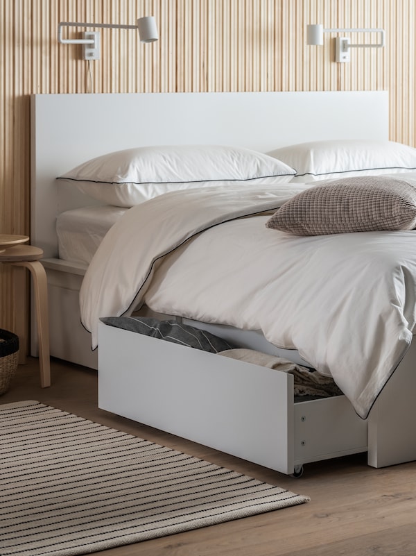 A white MALM bed with 4 storage boxes and KUNGSBLOMMA bed linen stands in a bedroom. One storage box is pulled out.