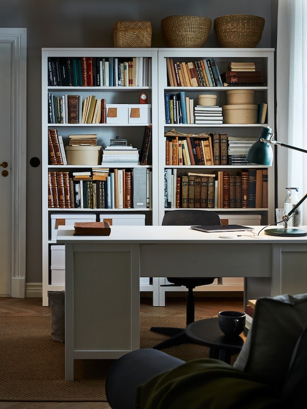 A workspace at home with a white HEMNES desk and matching bookcases placed behind it with books, boxes and baskets.