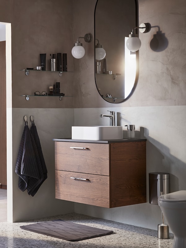 A contemporary bathroom with two FRIHULT wall lamps on either side of a LINDBYN mirror, above a wash-stand with two drawers.