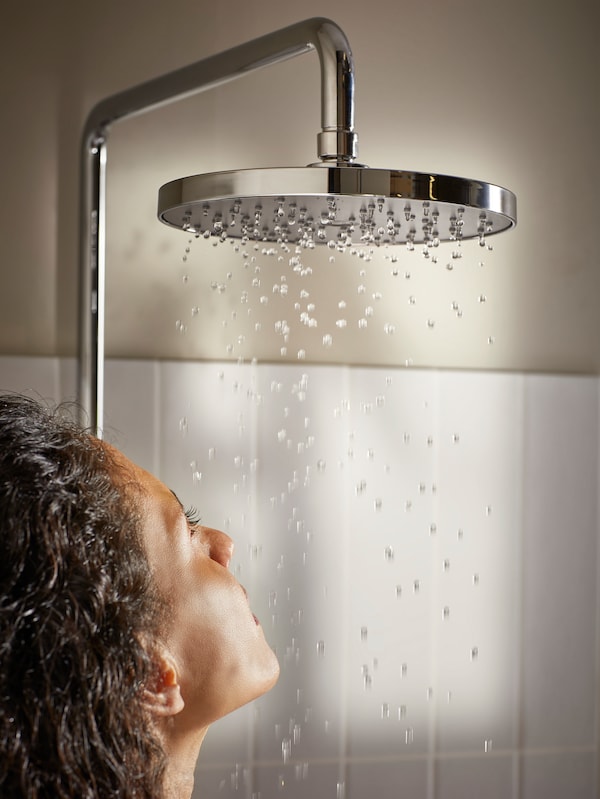 A bathroom with white tiles and beige walls. A woman is standing underneath a BROGRUND head/hand shower kit, taking a shower.