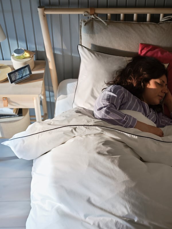 A person asleep in a birch BJÖRKSNÄS bed with white bed linen placed in front of a grey-blue wood panelled wall.