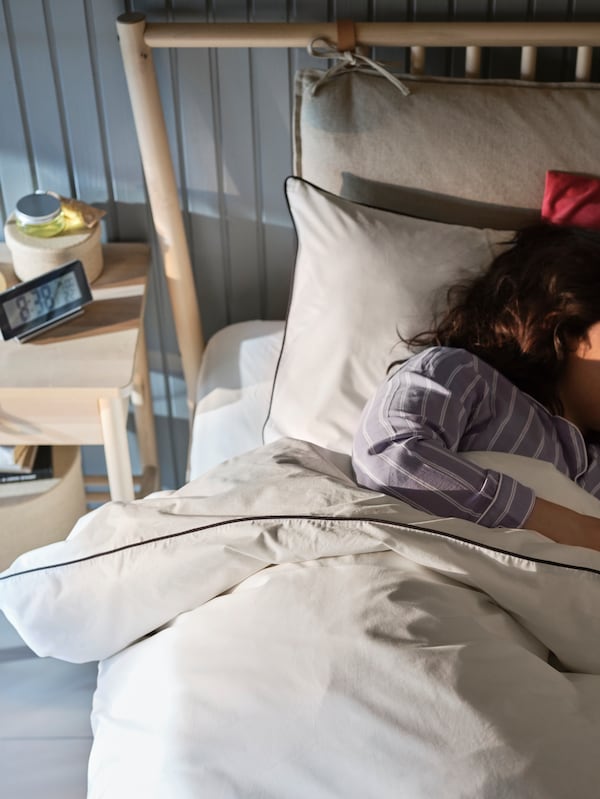 A person asleep in a birch BJÖRKSNÄS bed with white KUNGSBLOMMA bed linen placed in front of a grey-blue wood panelled wall.