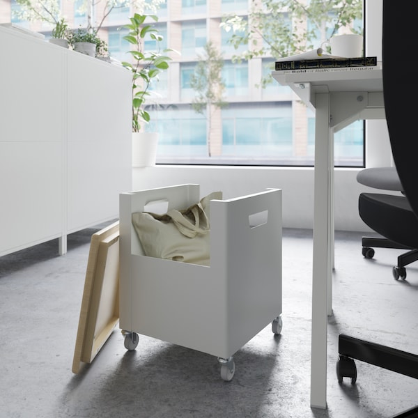 A white TROTTEN storage unit on casters with the top off and a beige bag inside, beside a table and an office chair.