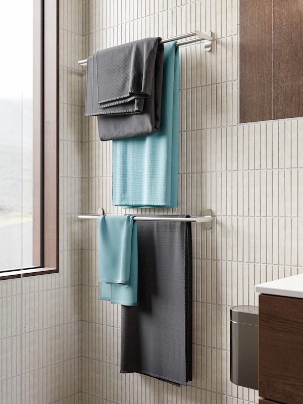A beige tiled bathroom, SALVIKEN bath towels in blue and anthracite hang from two BROGRUND towel rails in stainless steel.
