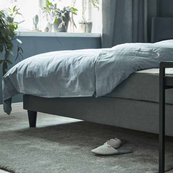 A grey SÄBÖVIK divan bed with KOPPARBLAD bed linen stands near a window. Under the bed is a rug with a pair of slippers.