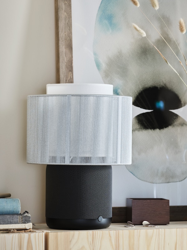 A lamp consisting of a black SYMFONISK speaker lamp base with WiFi and a white shade stands in front of a picture on a wall.