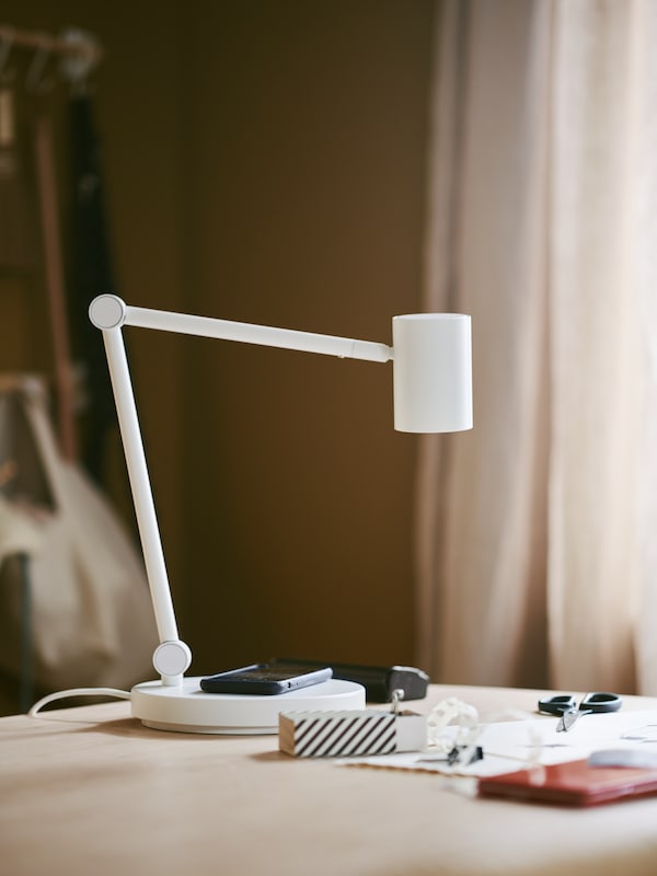 A mobile phone lies on top of the base of a NYMÅNE work lamp with wireless charging which is standing on a table.