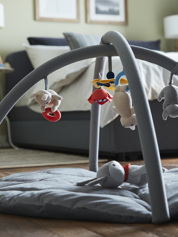 A GULLIGAST soft toy shaped like a rabbit lies in a multicolor GULLIGAST baby gym near a grey HAUGA upholstered bed.