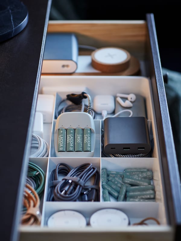 A pulled-out drawer revealing a white KUGGIS eight-compartment insert filled with batteries, chargers and cables.