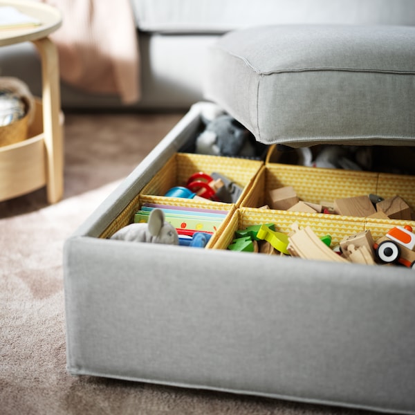 A light-grey KIVIK footstool with its lid half-off, revealing toys sorted in multiple yellow UPPRYMD boxes.