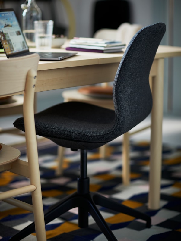 A dark grey LÅNGFJÄLL swivel chair stands by a RÖNNINGE birch table with a laptop, notebooks, and a glass of water on it.