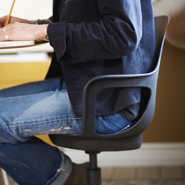 A person with a blue shirt and blue jeans sits on an anthracite ODGER swivel chair by a table.