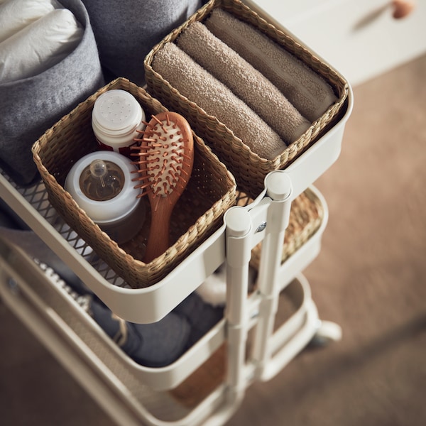 A white RÅSKOG cart holding LURPASSA seagrass boxes that in turn are filled with textiles and accessories.