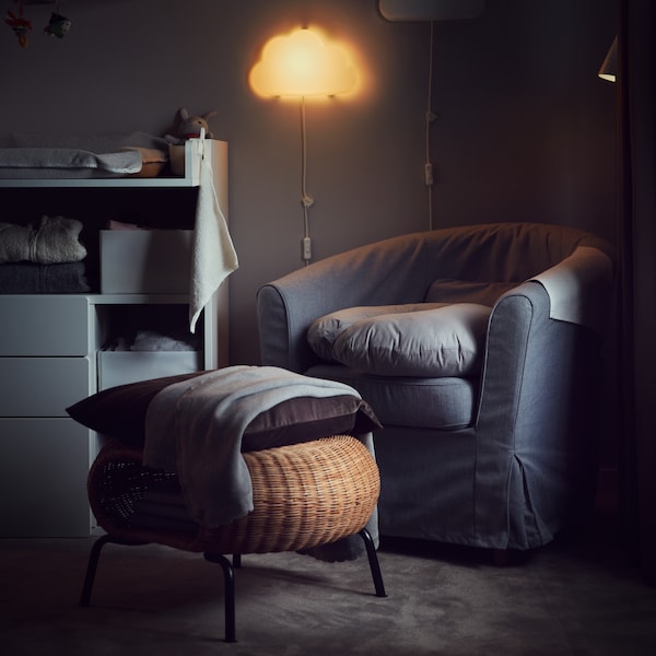 A cushion on a rattan GAMLEHULT footstool with storage in front of an armchair in a room lit by a night wall lamp.