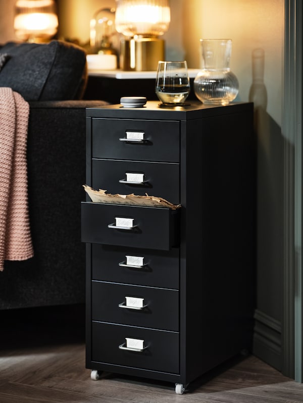 A black HELMER drawer unit on casters stands at an angle next to a sofa. Two lit brass lamps are on a shelf behind the sofa.