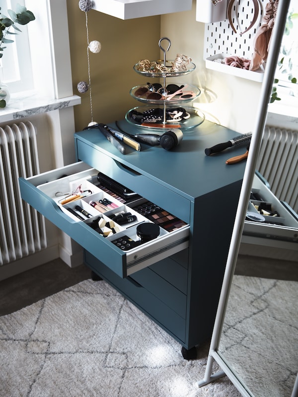 A grey-turquoise ALEX drawer unit with a drawer open and hair styling items inside and on top, plus a white standing mirror.