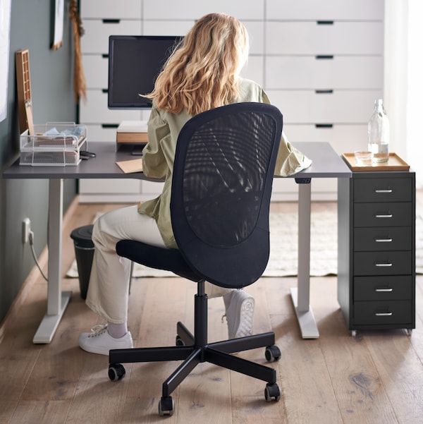 A woman sitting on a black FLINTAN office chair by a grey sit/stand RODULF desk with a computer, and a drawer unit.