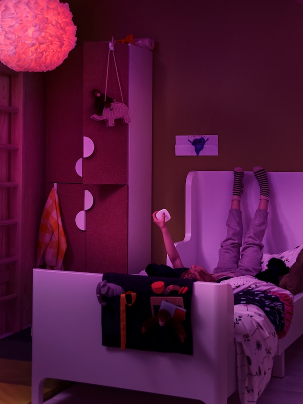 A little girl lying in her bed uses a STYRBAR remote to change the color of the LED bulb in her VINDKAST pendant lamp.