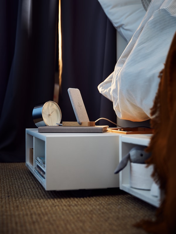 A FREDVANG underbed storage/bedside table, with a clock and other items on top, stands on a floor, partly underneath a bed.