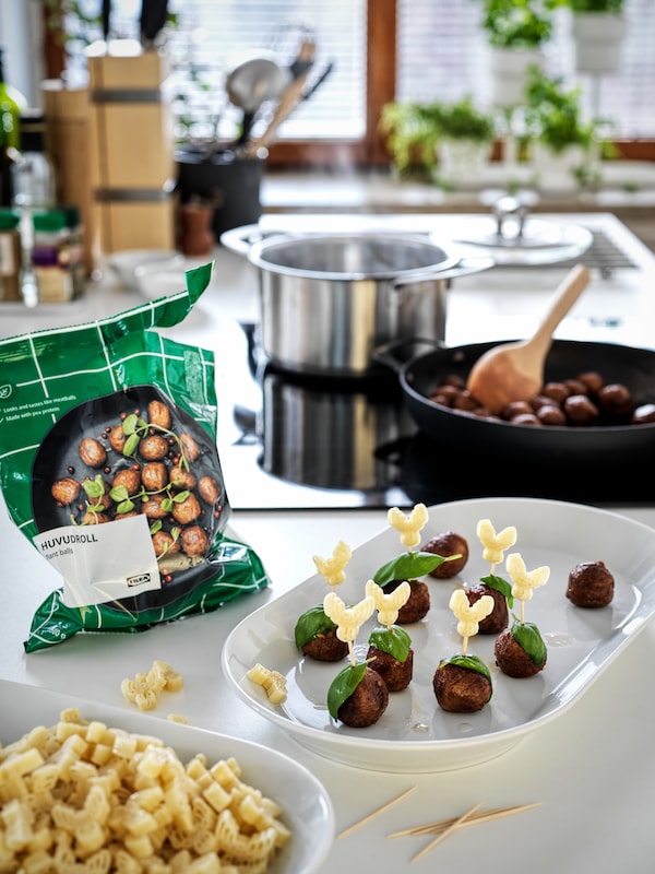 A bag of HUVUDROLL plant balls beside a plate of vegan meatball appetisers; a frying pan contains more in the back.
