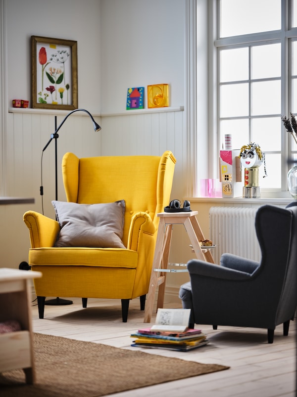 A living room corner with a yellow STRANDMON wing chair and a grey STRANDMON children’s armchair.