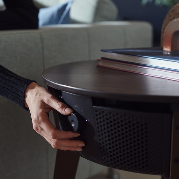 A woman’s hand adjusts a control knob on a black STARKVIND table with air purifier which is standing next to a sofa.