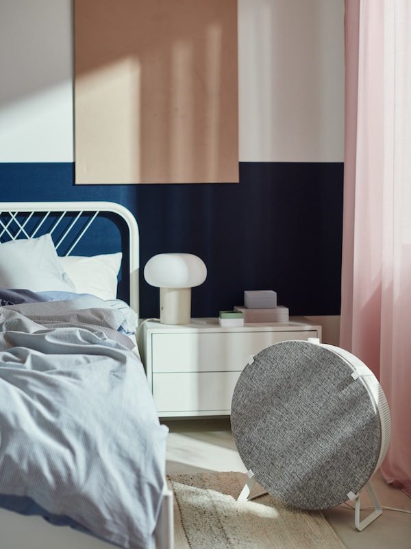 In a bedroom, a white STARKVIND air purifier stands on the floor at the side of a white NESTTUN bed.