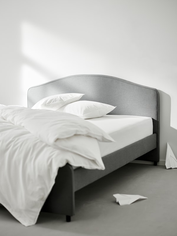 A gray HAUGA upholstered bed with a duvet and two pillows in ÄNGSLILJA bed linen. Two paper planes are on the floor nearby.