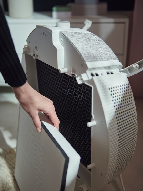 A woman’s hand removes a particle filter from the front of a white STARKVIND air purifier which is standing on the floor.